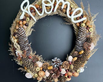 May include: A circular wreath with a variety of natural elements, including pinecones, acorns, and dried grasses. The word "home" is spelled out in white yarn across the top. The wreath is set against a black background.