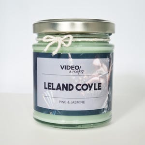 Pode incluir: Uma vela verde num frasco de vidro com tampa prateada. O rótulo diz "VIDEO wicks" e "LELAND COYLE PINE & JASMINE". Uma corda branca está amarrada ao redor da parte superior do frasco.