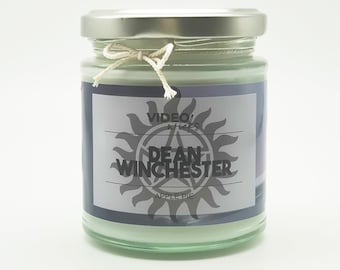 May include: A glass jar candle with a silver lid and a white label. The label has a black and white image of a sun with a pentagram inside. The text on the label reads "VIDEO/s" and "DEAN WINCHESTER" with "APPLE PIE" underneath.