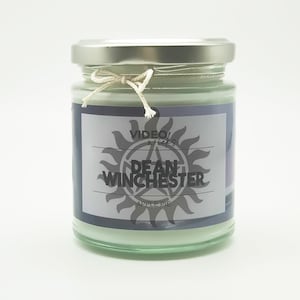 Puede incluir: Una vela en un tarro de vidrio con tapa plateada y una etiqueta blanca. La etiqueta tiene una imagen en blanco y negro de un sol con un pentagrama en su interior. El texto de la etiqueta dice "VIDEO/s" y "DEAN WINCHESTER" con "APPLE PIE" debajo.