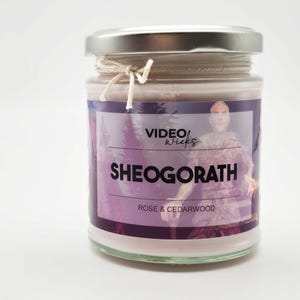 Puede incluir: Un tarro de vidrio para velas con tapa plateada y una etiqueta blanca. La etiqueta tiene un fondo morado y blanco con una imagen de un bosque y una persona. El texto de la etiqueta dice "VIDEO WICKS SHEOGORATH ROSE & CEDARWOOD".