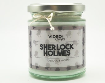 May include: A glass jar candle with a silver lid and a white label. The label has a black and white silhouette pattern and the text "VIDEO Wicks SHERLOCK HOLMES TOBACCO & WOOD".