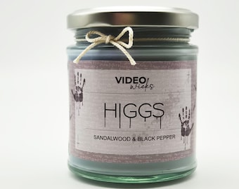 May include: A glass candle jar with a silver lid and a label that reads "VIDEO wicks HIGGS SANDALWOOD & BLACK PEPPER". The label has a distressed look with handprint graphics.