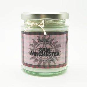 May include: A glass jar candle with a silver lid and a white label with red and black checked pattern. The label has the text "VIDEO WICKS" and "SAM WINCHESTER" in black text. The label also has a black and white symbol of a sun with a pentagram inside. The text "COFFEE & CARAMEL" is also on the label.