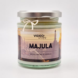 May include: A glass jar candle with a silver lid and a white string tied around the lid. The label on the jar says "VIDEO wicks" and "MAJULA WOOD SMOKE & VANILLA".