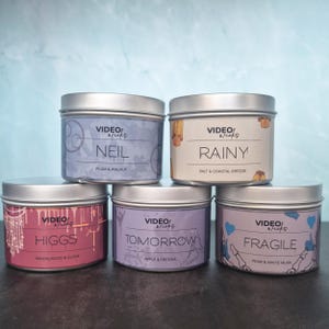 May include: Five scented candles in silver tins. Each tin has a label with a name and scent description: Neil (Plum & Walnut), Rainy (Salt & Coastal Breeze), Higgs (Sandalwood & Clove), Tomorrow (Apple & Freesia), and Fragile (Pear & White Musk).