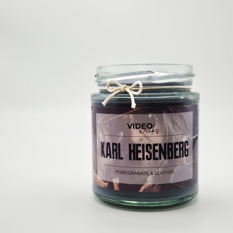 Puede incluir: Una vela en un tarro de vidrio con una etiqueta negra que dice "VIDEO/Wicks KARL HEISENBERG POMEGRANATE & LEATHER". La etiqueta tiene una imagen de una persona con cabello largo y oscuro.