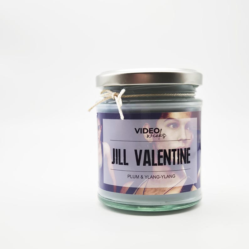 May include: A gray glass candle jar with a silver lid. The label reads "VIDEO WICKS JILL VALENTINE PLUM & YLANG-YLANG".