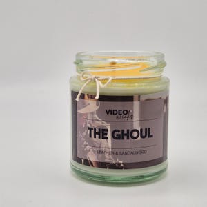 May include: A glass jar candle with a green label that says "VIDEO WICKS THE GHOUL LEATHER & SANDALWOOD". The label has a vintage photo of a person wearing a cowboy hat.