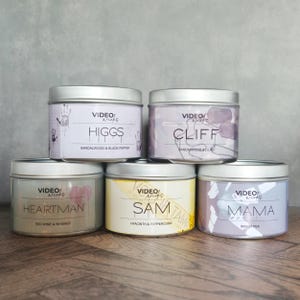 Puede incluir: Cinco latas de velas plateadas con etiquetas de diferentes colores. Las etiquetas tienen el texto "VIDEO/Wieks" y los nombres "HIGGS", "CLIFF", "HEARTMAN", "SAM" y "MAMA" con aromas correspondientes.