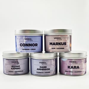 May include: Five silver candle jars with labels featuring different names and scents. The scents are Lavender + Cedar, Aloe + Cucumber, Jasmine + Cedarwood, Magnolia + Woods, and Pink Topaz + Magnolia.