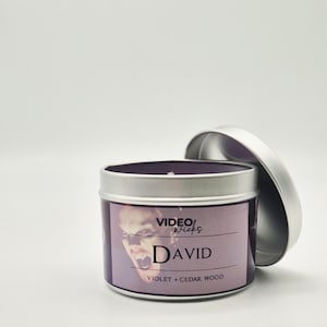 Peut inclure: Une bo&icirc;te de bougie en argent avec une &eacute;tiquette violette. L'&eacute;tiquette pr&eacute;sente une image en noir et blanc d'un visage d'homme avec le texte "VIDEO/wicks DAVID VIOLET + CEDAR WOOD".