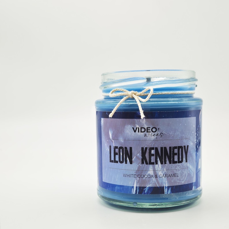 May include: A blue candle in a glass jar with a lid. The candle has a label that reads "LEON KENNEDY" and "WHITE COCOA & CARAMEL". A white string is tied around the top of the jar. The brand name "VIDEO wicks" is also visible.