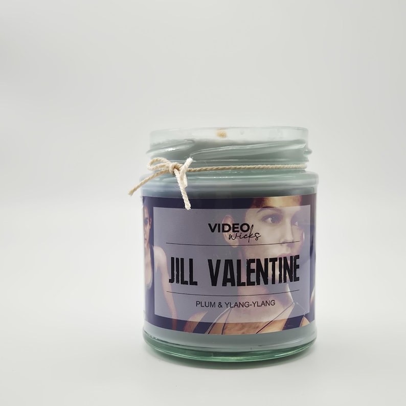 May include: A green glass candle jar with a white lid and a label that reads "VIDEO WICKS JILL VALENTINE PLUM & YLANG-YLANG". The label features a photo of a woman.