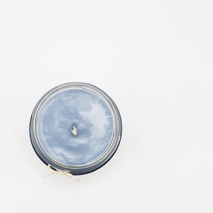 May include: Overhead view of a blue candle in a clear glass jar. The candle has a white wick and is tied with a natural twine bow. The candle is set against a white background.