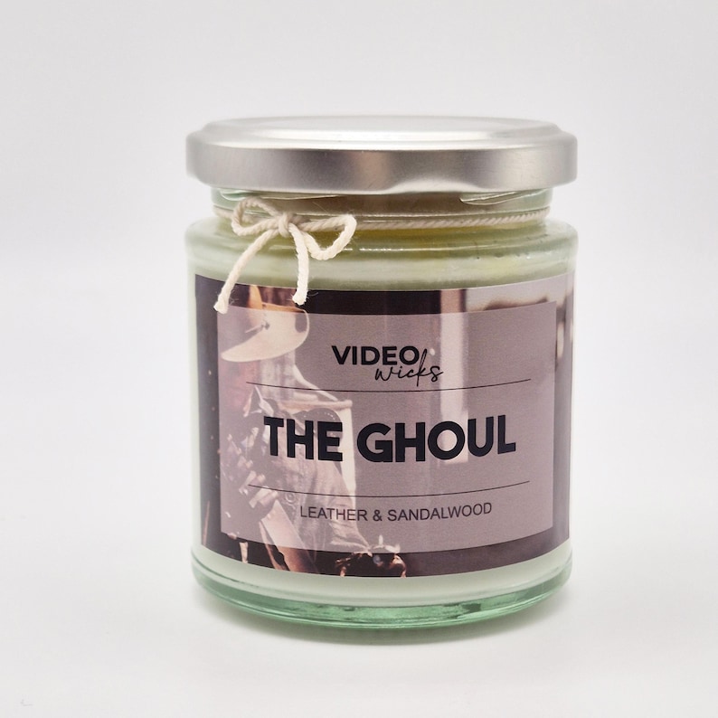 May include: A glass candle jar with a silver lid and a white string bow. The label reads "VIDEO WICKS THE GHOUL LEATHER & SANDALWOOD".