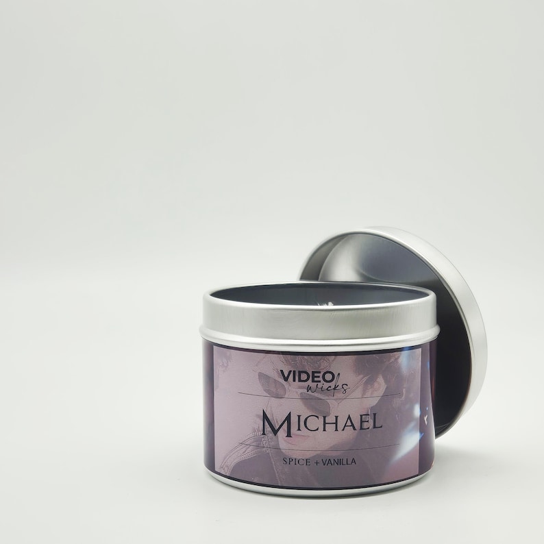 Peut inclure: Une bo&icirc;te de bougie en argent avec un couvercle noir. L'&eacute;tiquette indique "VIDEO wicks MICHAEL SPICE + VANILLA". L'&eacute;tiquette pr&eacute;sente un motif violet et blanc avec le visage d'une personne.