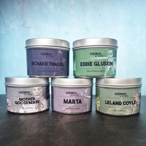 May include: Five metal tin candles with lids. Each candle has a different label with a name and scent description. The labels are in shades of purple, green, and grey. The text "Video Wicks" is at the top of each label.