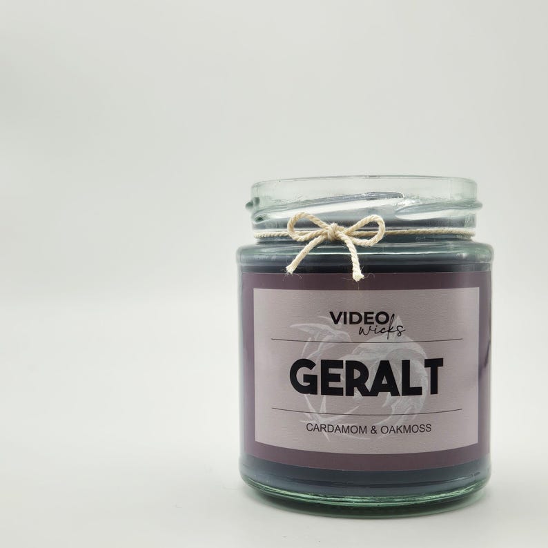 May include: A glass jar candle with a black label that reads "VIDEO WICKS GERALT CARDAMOM & OAKMOSS". The jar has a natural twine bow around the lid.