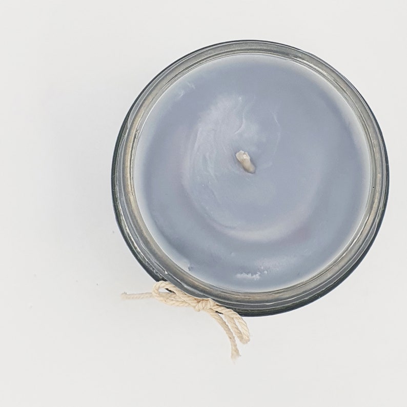 May include: A gray candle in a glass jar with a white wick. The candle has a light gray color and a smooth surface. The jar has a clear glass exterior and a black lid.