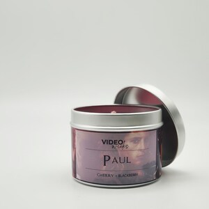 Peut inclure: Une bo&icirc;te de bougie en argent avec un couvercle rouge. L'&eacute;tiquette pr&eacute;sente une photo d'un homme avec le texte "VIDEO WICKS PAUL CHERRY + BLACKBERRY".