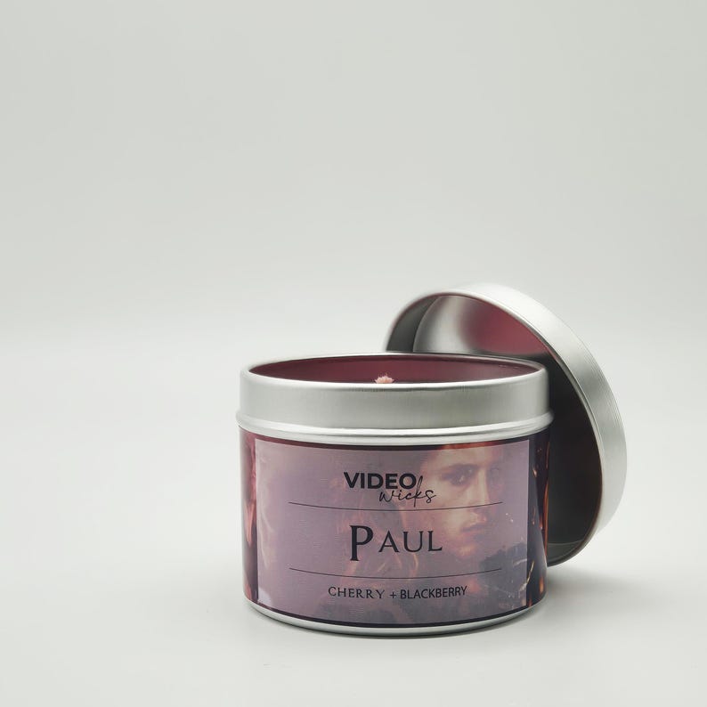 Peut inclure: Une bo&icirc;te de bougie en argent avec un couvercle rouge. L'&eacute;tiquette pr&eacute;sente une photo d'un homme avec le texte "VIDEO WICKS PAUL CHERRY + BLACKBERRY".