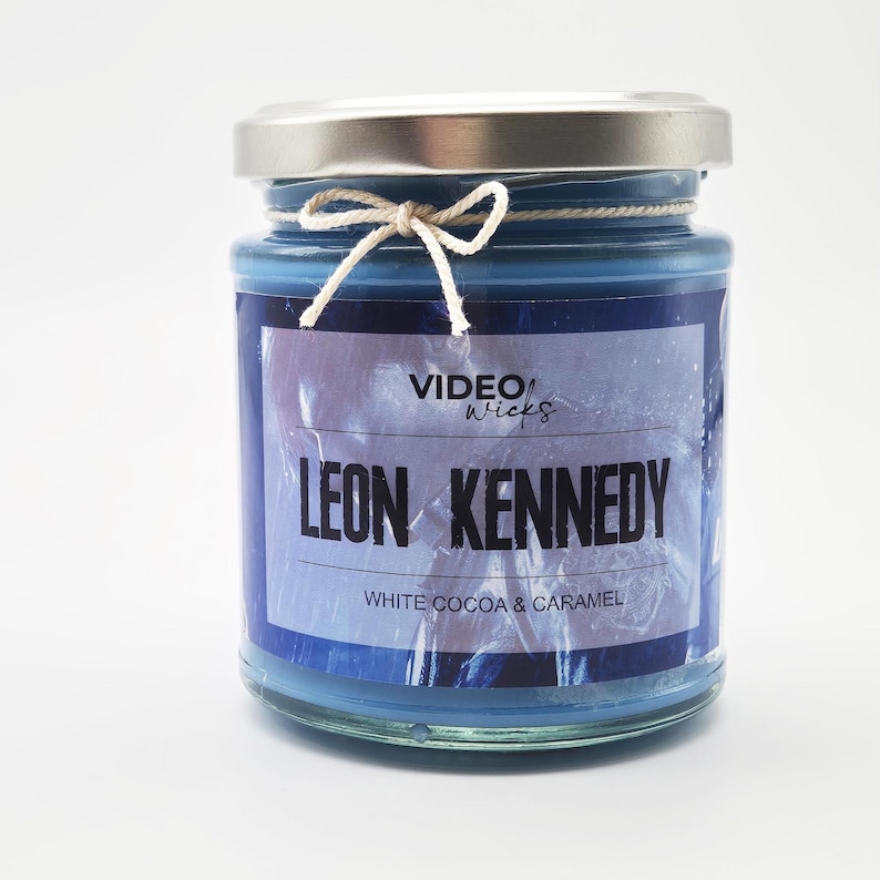 May include: A blue glass jar candle with a silver lid and a decorative string bow. The label reads "VIDEO Wicks" and "LEON KENNEDY" with "WHITE COCOA & CARAMEL" below. The candle is a home decor item.