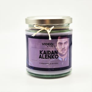 May include: A glass jar candle with a purple label featuring a man's face and the text "VIDEO wicks  KAIDAN ALENKO CINNAMON & MYRRH".