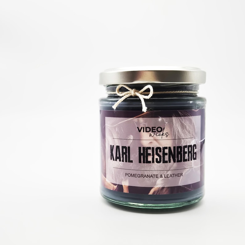 Puede incluir: Un tarro de vela de vidrio negro con tapa plateada y una etiqueta blanca. La etiqueta dice "VIDEO WICKS" y "KARL HEISENBERG" en texto negro. La etiqueta tambi&eacute;n dice "POMEGRANATE & LEATHER" en texto negro m&aacute;s peque&ntilde;o.