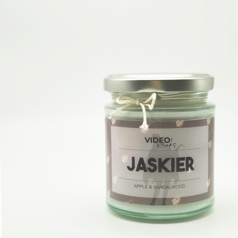 Op de afbeelding: Een glazen potkaars met een zilveren deksel en een wit etiket. Het etiket heeft de tekst "VIDEO WICKS" en "JASKIER" in zwarte tekst. Het etiket heeft ook de tekst "APPLE & SANDALWOOD" in kleinere zwarte tekst.