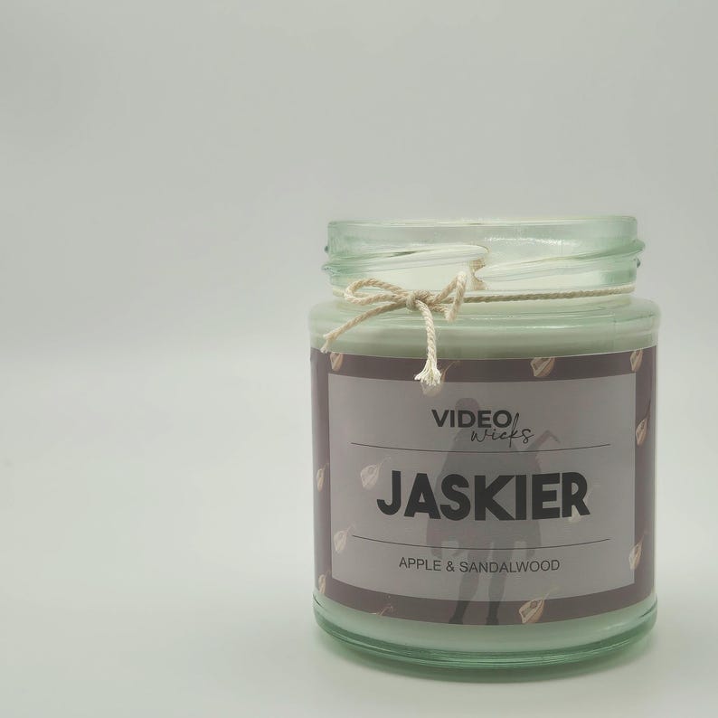 Op de afbeelding: Een glazen potkaars met een bruin etiket waarop "VIDEO Wicks" en "JASKIER" in witte tekst staat. Het etiket vermeldt ook "APPLE & SANDALWOOD" in witte tekst. De kaars is vastgebonden met een natuurlijke touw strik.