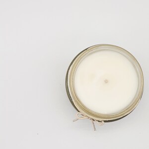 Op de afbeelding: Een witte kaars in een glazen pot met een bruine touwtjeslus. De kaars is niet aangestoken en heeft een kleine inkeping in het midden.