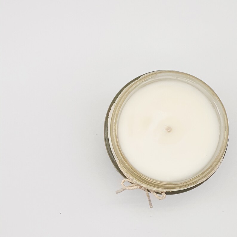 Op de afbeelding: Een witte kaars in een glazen pot met een bruine touwtjeslus. De kaars is niet aangestoken en heeft een kleine inkeping in het midden.