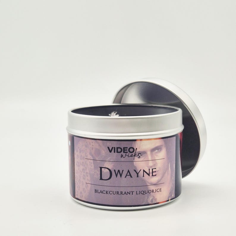 Peut inclure: Une bo&icirc;te de bougie en argent avec un couvercle noir. L'&eacute;tiquette pr&eacute;sente une photo d'un homme avec le texte "VIDEO WICKS" et "DWAYNE" en gras. Le texte "BLACKCURRANT LIQUORICE" est imprim&eacute; sous le nom.