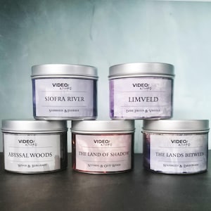 May include: Five silver-lidded candle tins with labels. The labels read "SIOFRA RIVER," "LIMVELD," "ABYSSAL WOODS," "THE LAND OF SHADOW," and "THE LANDS BETWEEN." Each label includes scent descriptions like "Seabreeze & Florals" and "Dark Fruits & Vanilla."