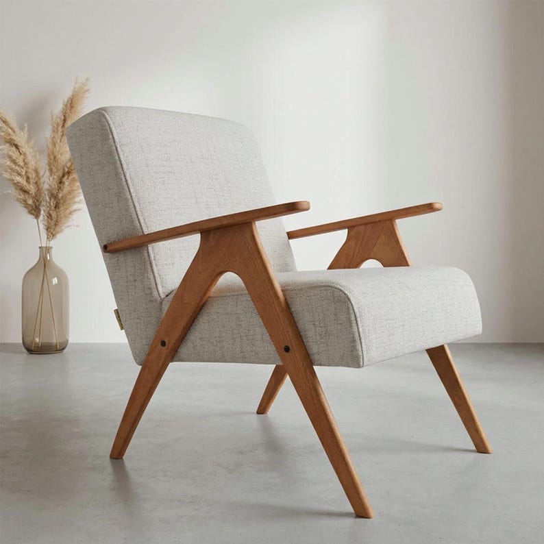 May include: A mid-century modern armchair with a light gray fabric seat and back. The chair features a solid wood frame with angled legs and armrests. The chair is designed for comfort and style.