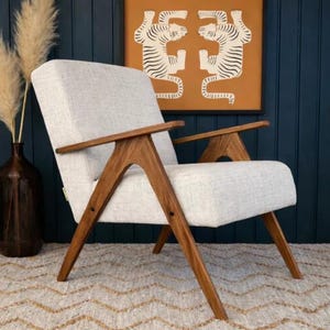May include: A mid-century modern armchair with a light gray upholstered seat and back. The chair features angled, solid wood armrests and legs. The background includes a framed tiger art print and a dark blue wall.