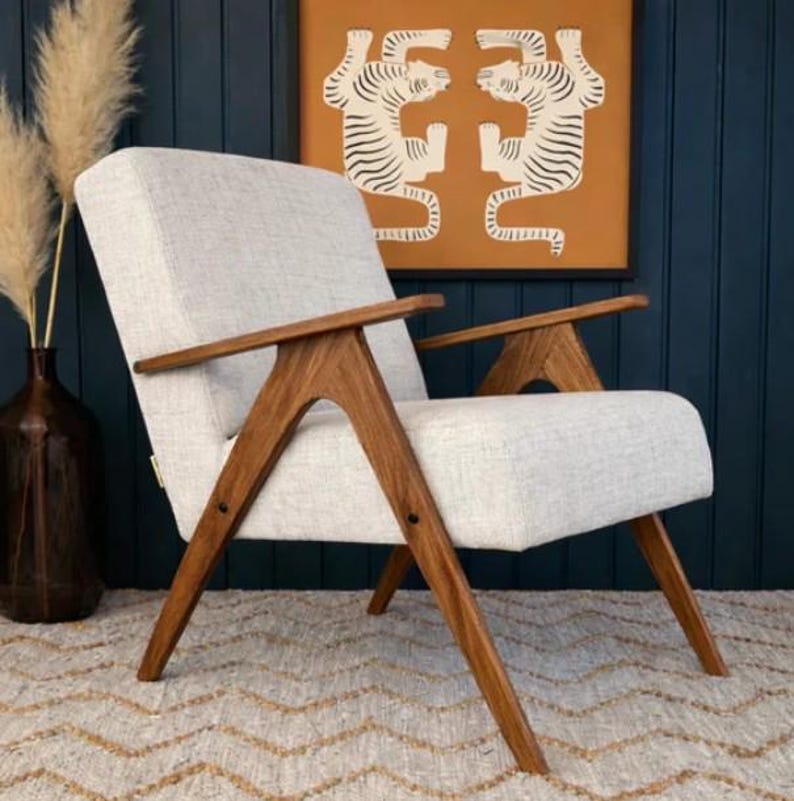 May include: A mid-century modern armchair with a light gray upholstered seat and back. The chair features angled, solid wood armrests and legs. The background includes a framed tiger art print and a dark blue wall.