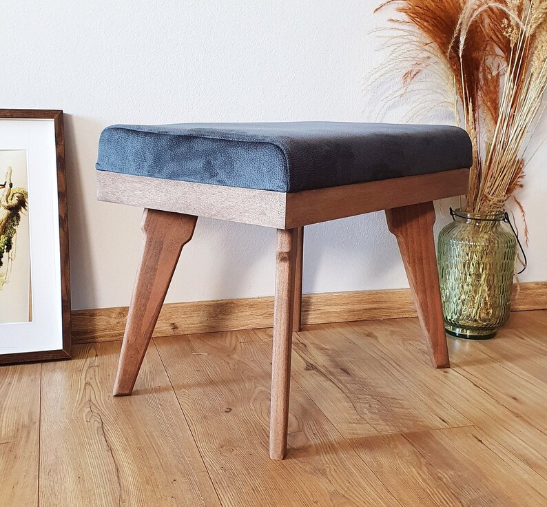 May include: A dark gray upholstered ottoman with a wooden frame and four legs. The ottoman is sitting on a wooden floor.