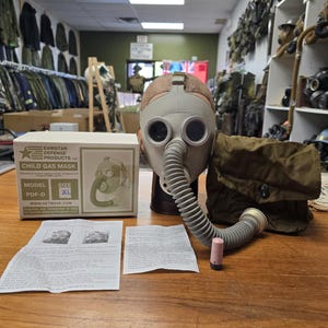 May include: A gray child gas mask with a corrugated hose and a matching olive green carrying bag. The mask is displayed with its original box and instruction sheets. The box reads "CHILD GAS MASK" and "MODEL PDF-D".