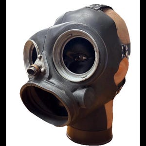 Puede incluir: Una máscara de gas negra con lentes oculares grandes y circulares y una boquilla prominente. La máscara está hecha de un material oscuro, posiblemente gomoso, con detalles metálicos alrededor de las lentes y una correa para sujetarla a la cabeza. La máscara está sobre una cabeza de maniquí.