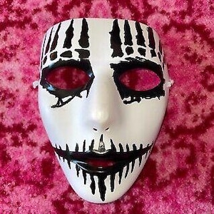 May include: A white mask with black accents, featuring a stitched mouth design and eye details. The mask has a smooth surface and is designed with a theatrical or costume theme. The eye holes are dark red.