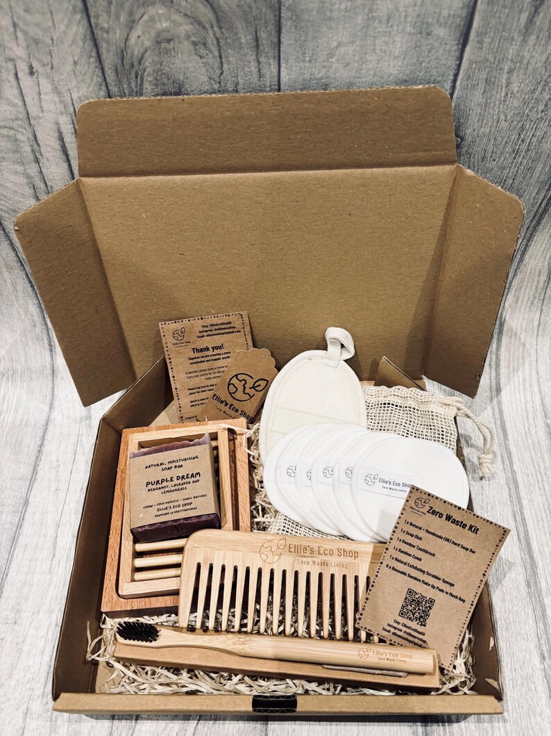 May include: An open cardboard box filled with eco-friendly zero waste products. Includes a soap bar, wooden comb, bamboo toothbrush, reusable cotton pads, and a small drawstring bag. The box contains a card that says "Zero Waste Kit".