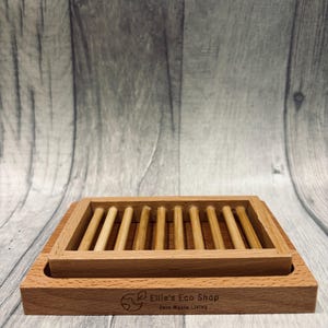 May include: A rectangular wooden soap dish with a slatted top and a lower tray. The dish is made of light brown wood and features the text "Ellie's Eco Shop Zero Waste Living".