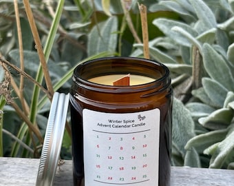 May include: A brown glass jar candle with a wooden wick and a white label that says "Winter Spice Advent Calendar Candle". The label also includes a calendar with numbers 1 through 25. The candle is hand-poured in Hertfordshire and made with beeswax.