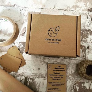 May include: A brown cardboard box with the logo "Ellie's Eco Shop" and the text "Zero Waste Living". The box is on a white and gray brick surface, with a roll of tape, tags, and twine.