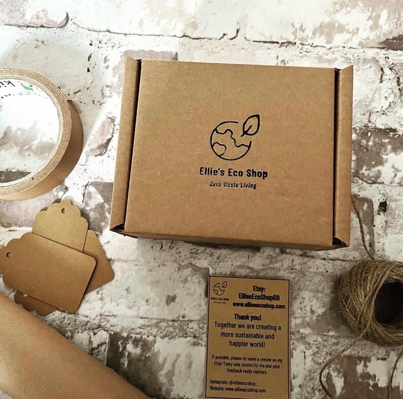 May include: A brown cardboard box with the logo "Ellie's Eco Shop" and the text "Zero Waste Living". The box is on a white and gray brick surface, with a roll of tape, tags, and twine.