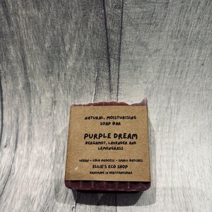 May include: A rectangular, dark purple soap bar with a brown paper label. The label reads "Purple Dream" with ingredients listed below. The soap is handmade and vegan. The background is a gray wooden surface.