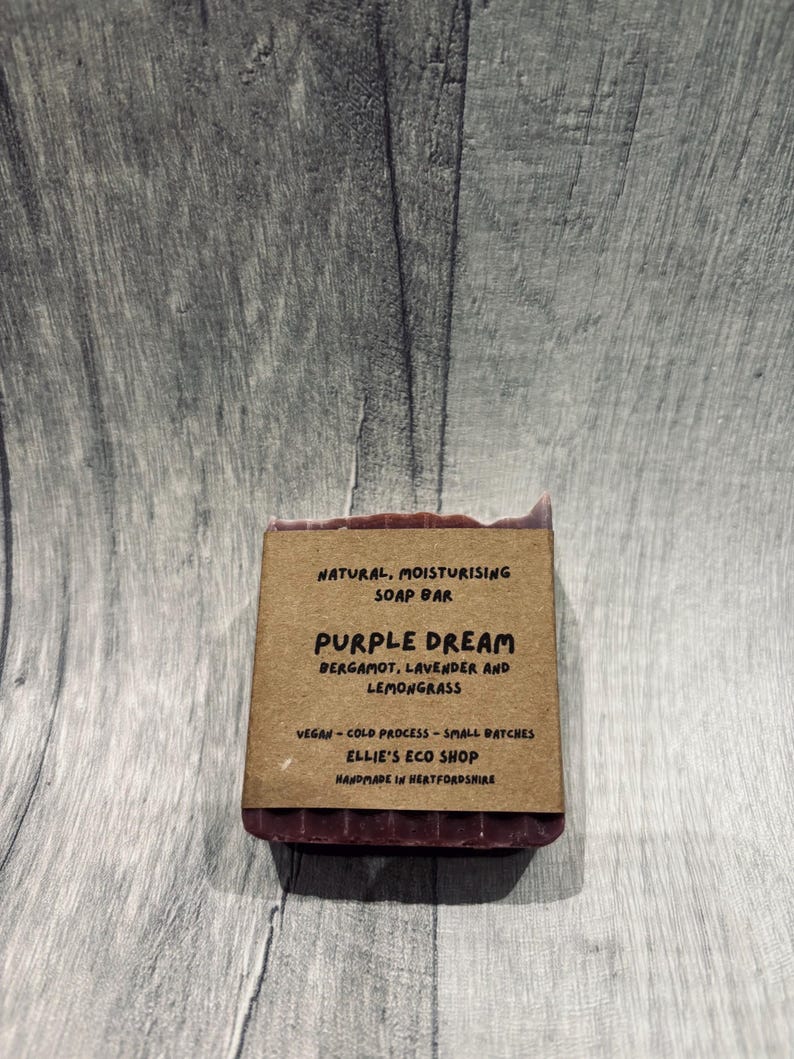 May include: A rectangular, dark purple soap bar with a brown paper label. The label reads "Purple Dream" with ingredients listed below. The soap is handmade and vegan. The background is a gray wooden surface.