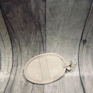 May include: An oval-shaped, beige bath sponge with a loop for hanging. The sponge has a textured surface and a vertical band across the center. The background is a gray wooden surface.